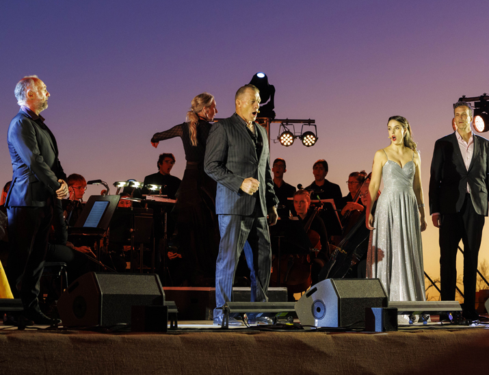 Opera singers performing on stage under open, outback sky. 